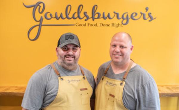 picture of two men in front of a yellow wall with the word gouldsburger written on it. 