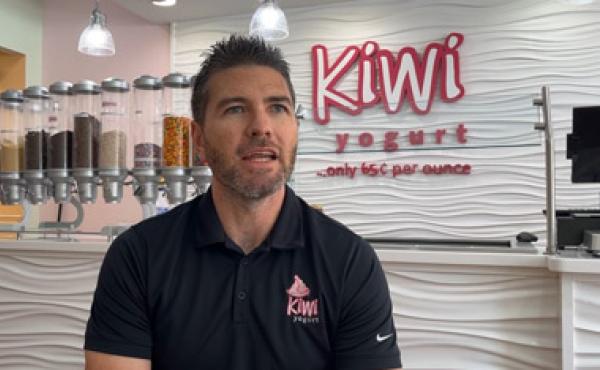 man in black polo sitting in front of a Kiwi yogurt sign on a wall