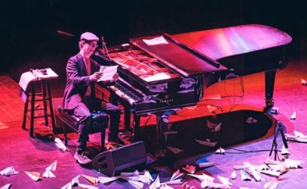 man sitting at a piano reading a paper with paper planes all around his feet