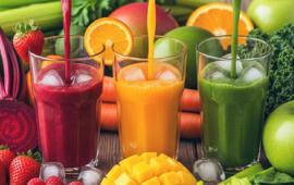 three glass cups surrounded by fresh fruit with red, yellow, and green juices being poared into each glass. 